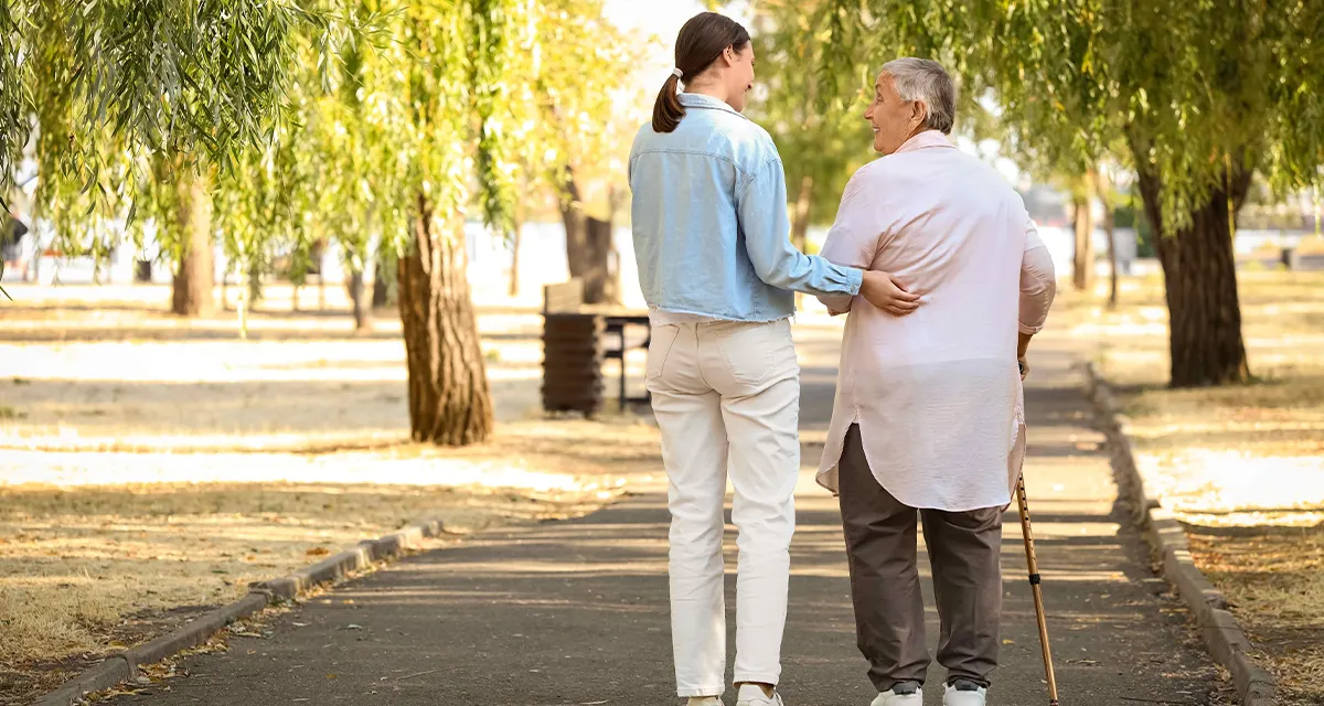 Une accompagnante soutient une personne âgée marchant avec une canne lors d'une promenade dans un parc
